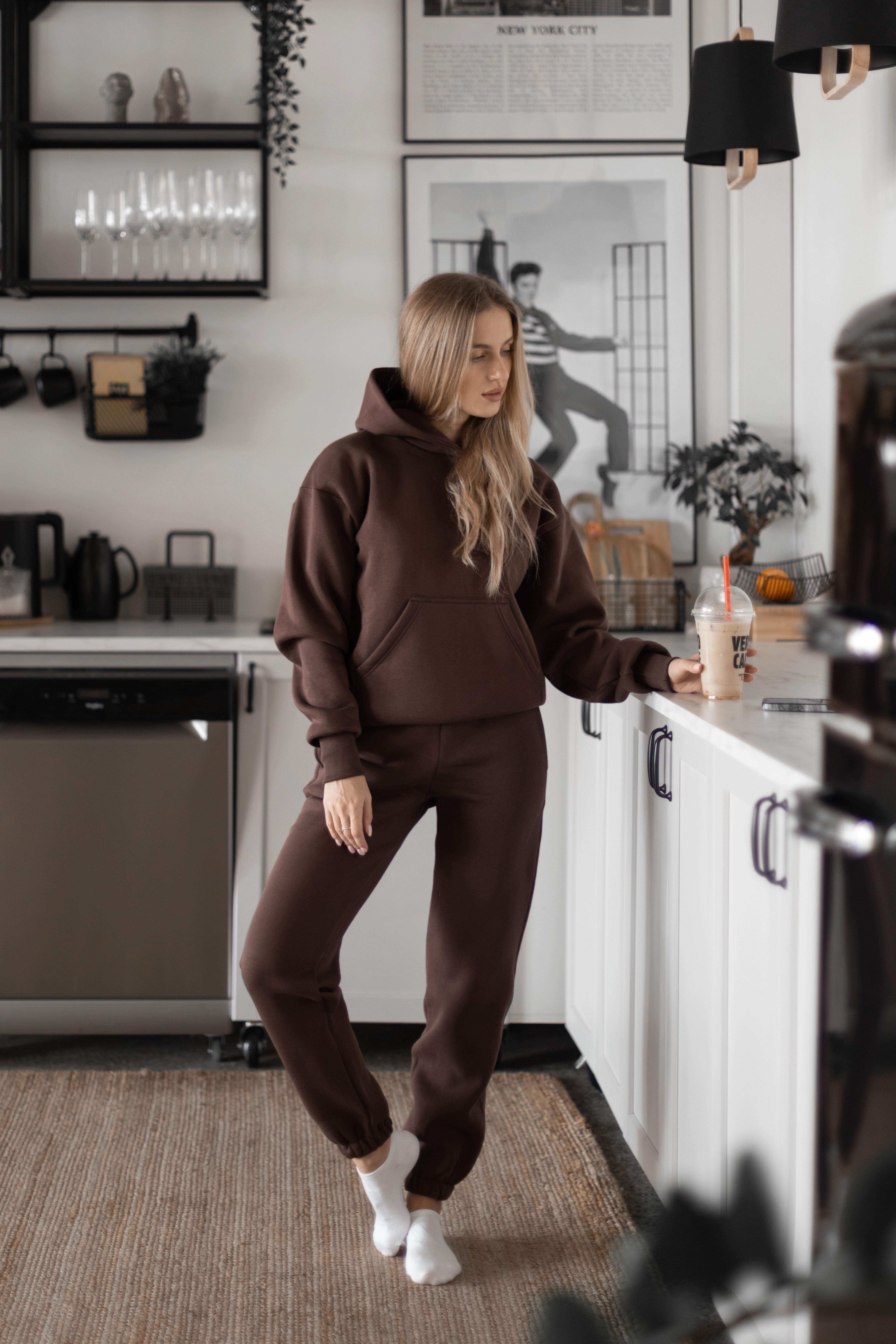 Moteris su tamsiai rudu treningu ir ilgu palaidais plaukais stovi virtuvėje, laiko gėrimą su šiaudeliu, matomas modernus interjeras