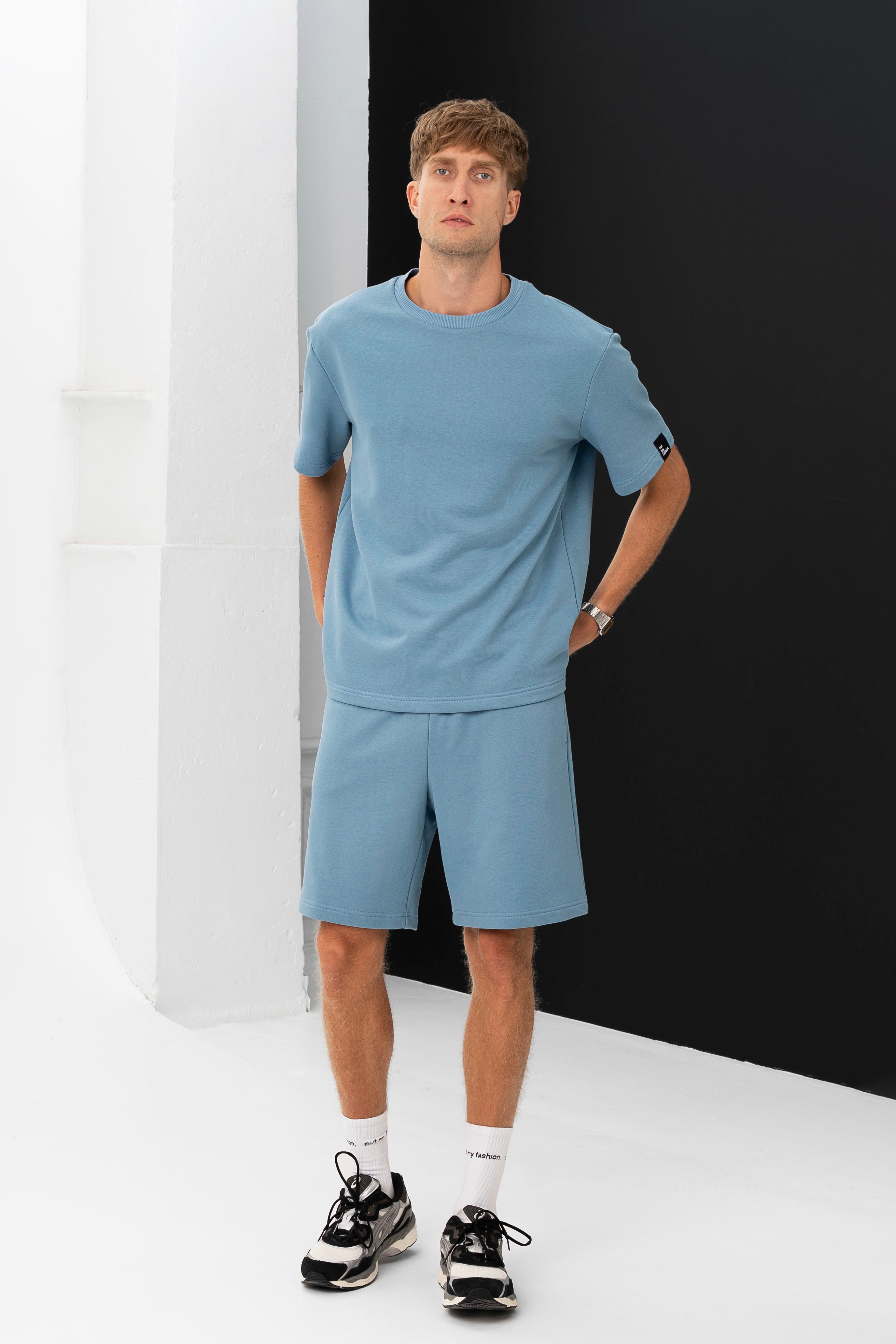 Vyriškas šviesiai mėlynas laisvalaikio kostiumas su šortais ir marškinėliais, modelis stovi žiūrėdamas į šoną