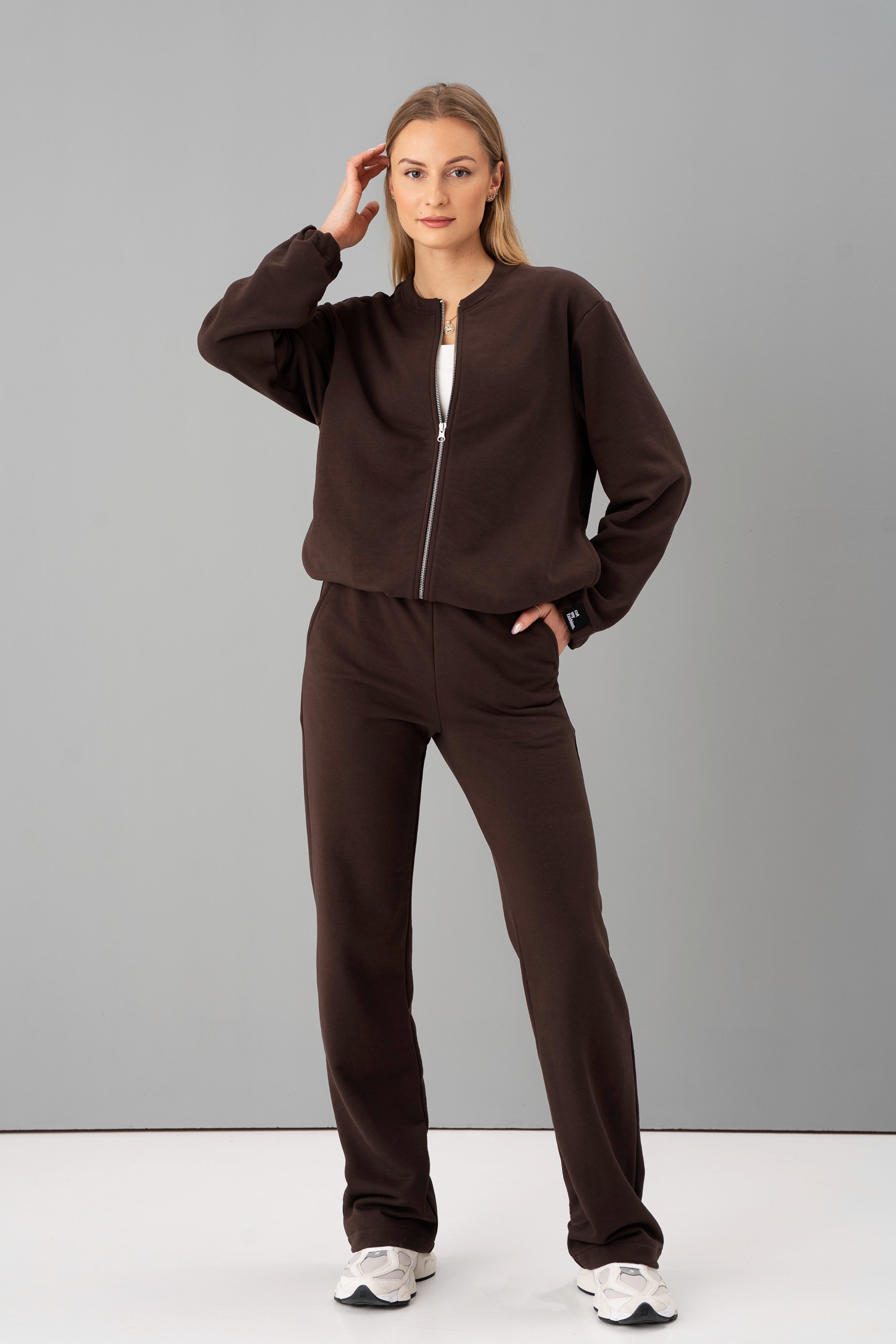 modelis su rudu treningų kostiumu mėja, rankas įkišusi į kišenes, pozuoja studijoje