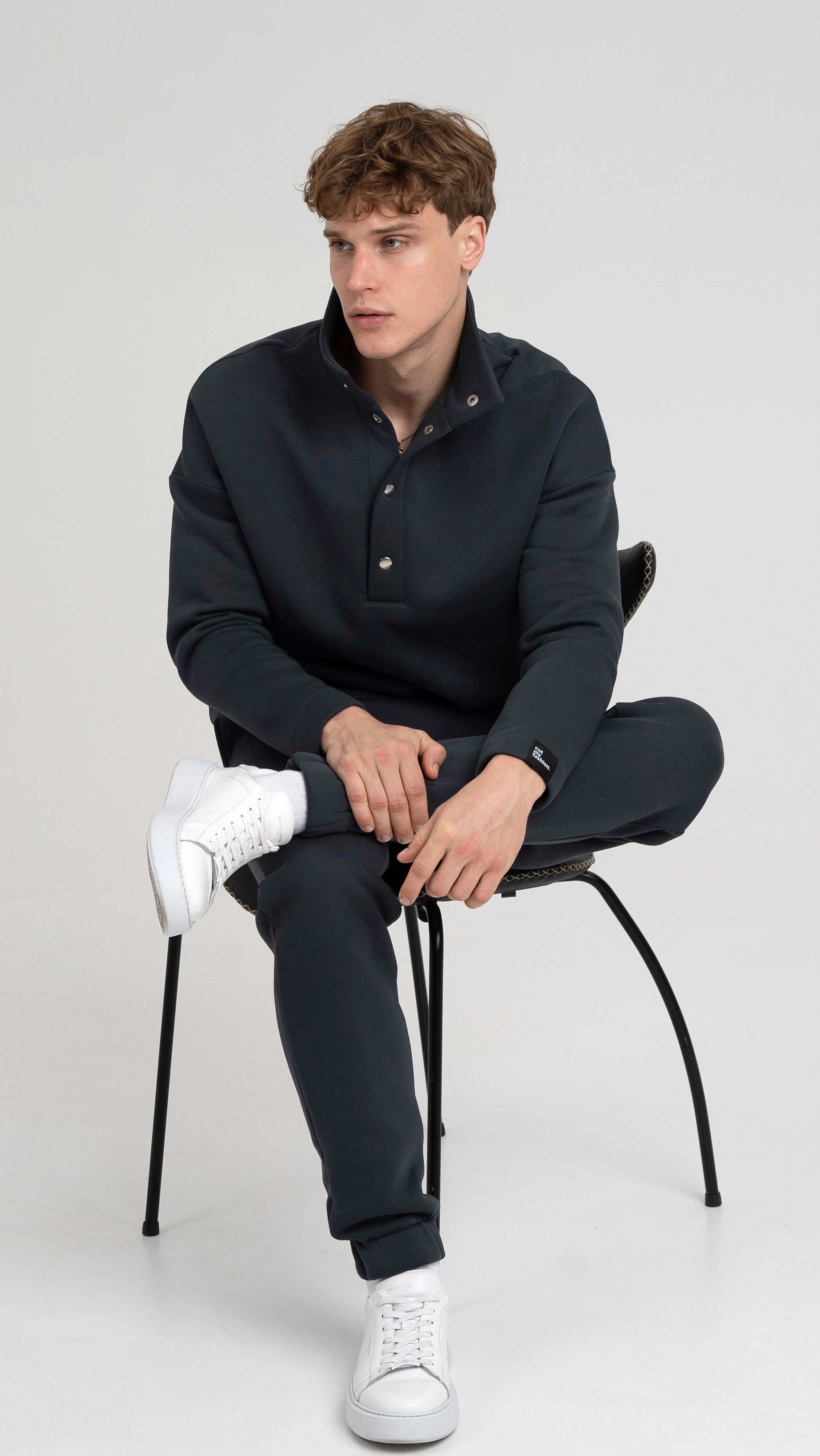 Modelis sėdi ant kėdės, vilkėdamas tamsiai pilkus treningus su sagomis, prie kurių dera balti sportbačiai