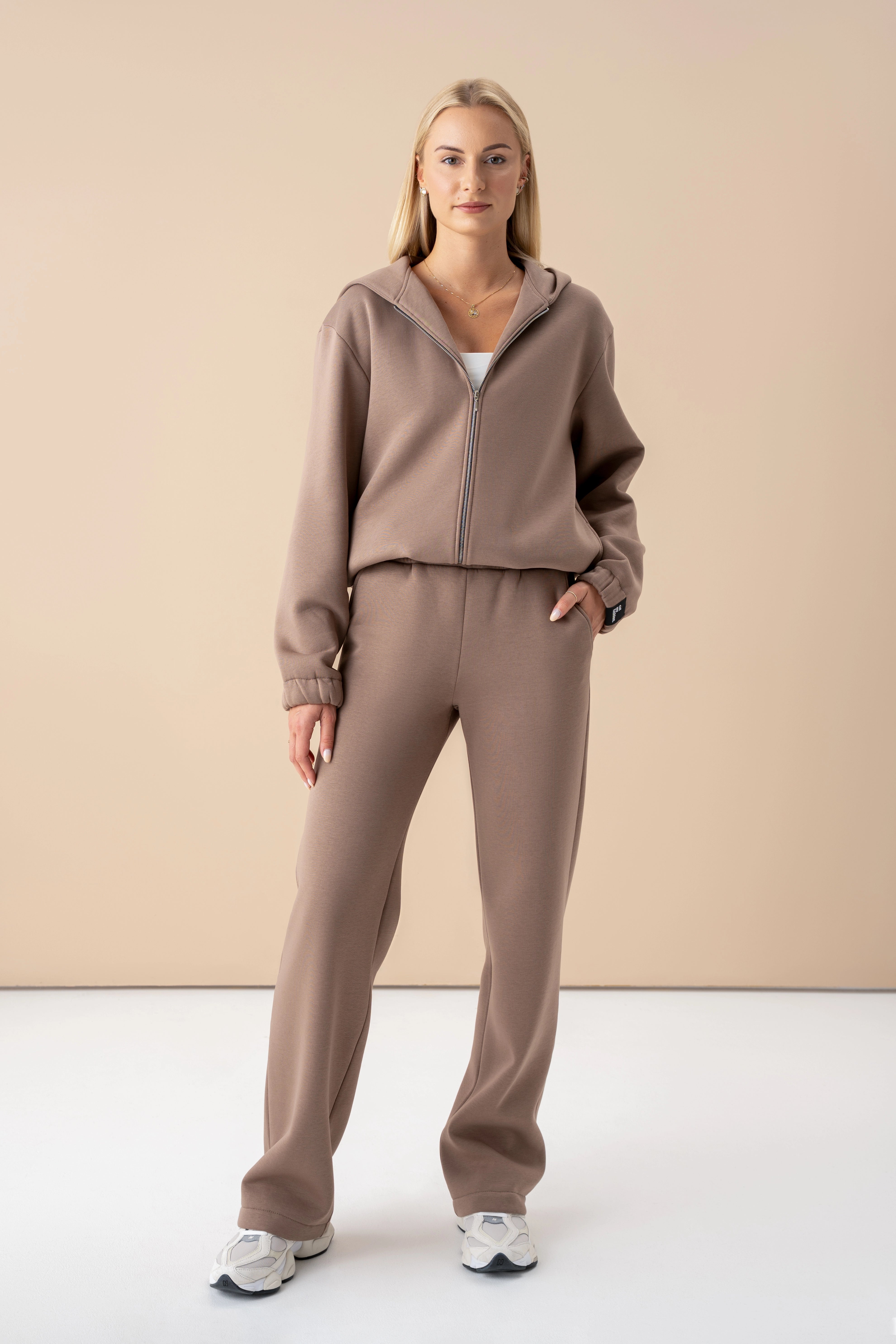 modelis dėvi latte spalvos laisvalaikio kostiumą pagamintą Lietuvoje, stovi studijoje, ranką įkišusi į kišenę, žiūri į objektyvą