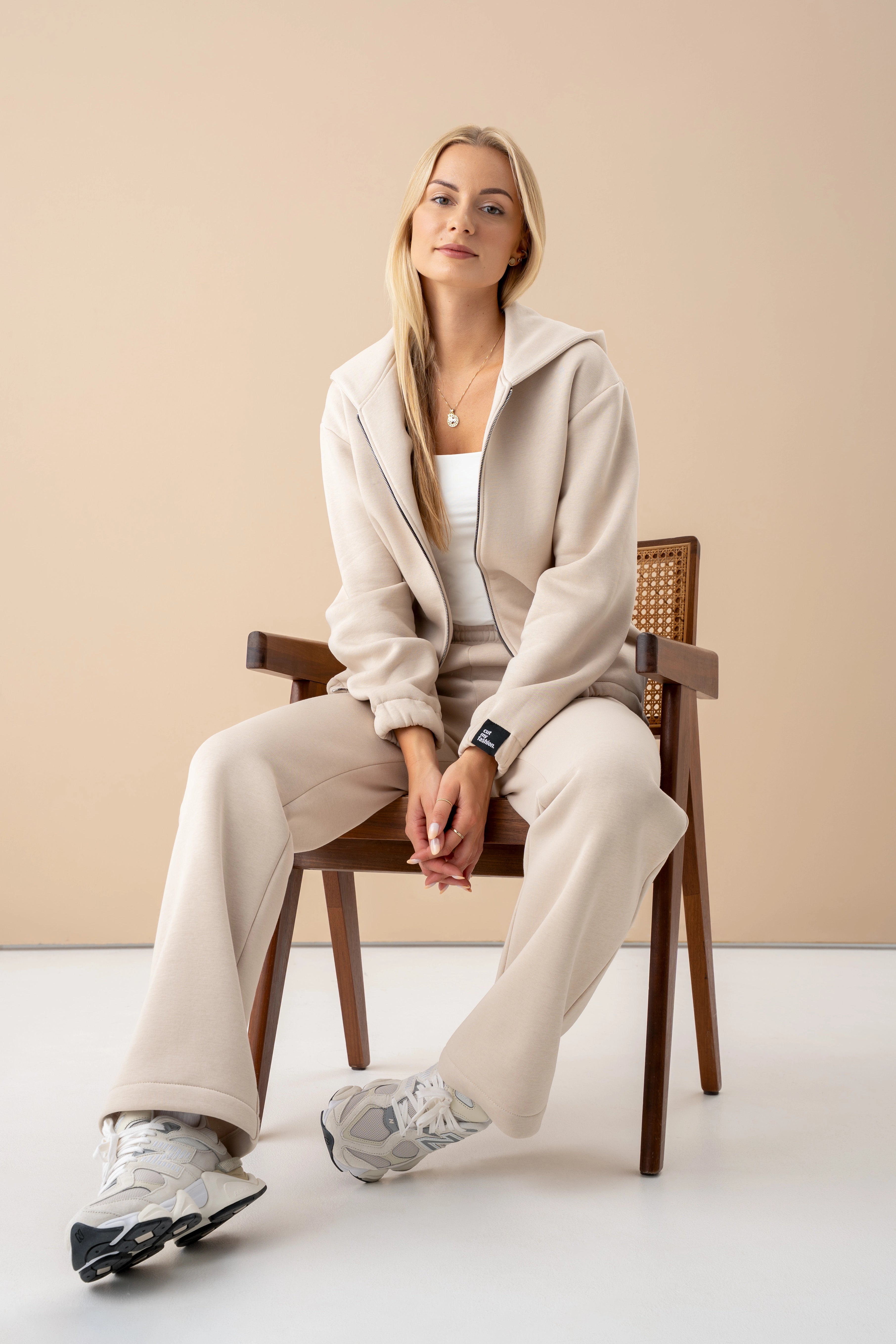 moteris su cutmyfashion treningais sėdi ant kėdės, dėvi užtraukiamą džemperį, žiūri į objektyvą