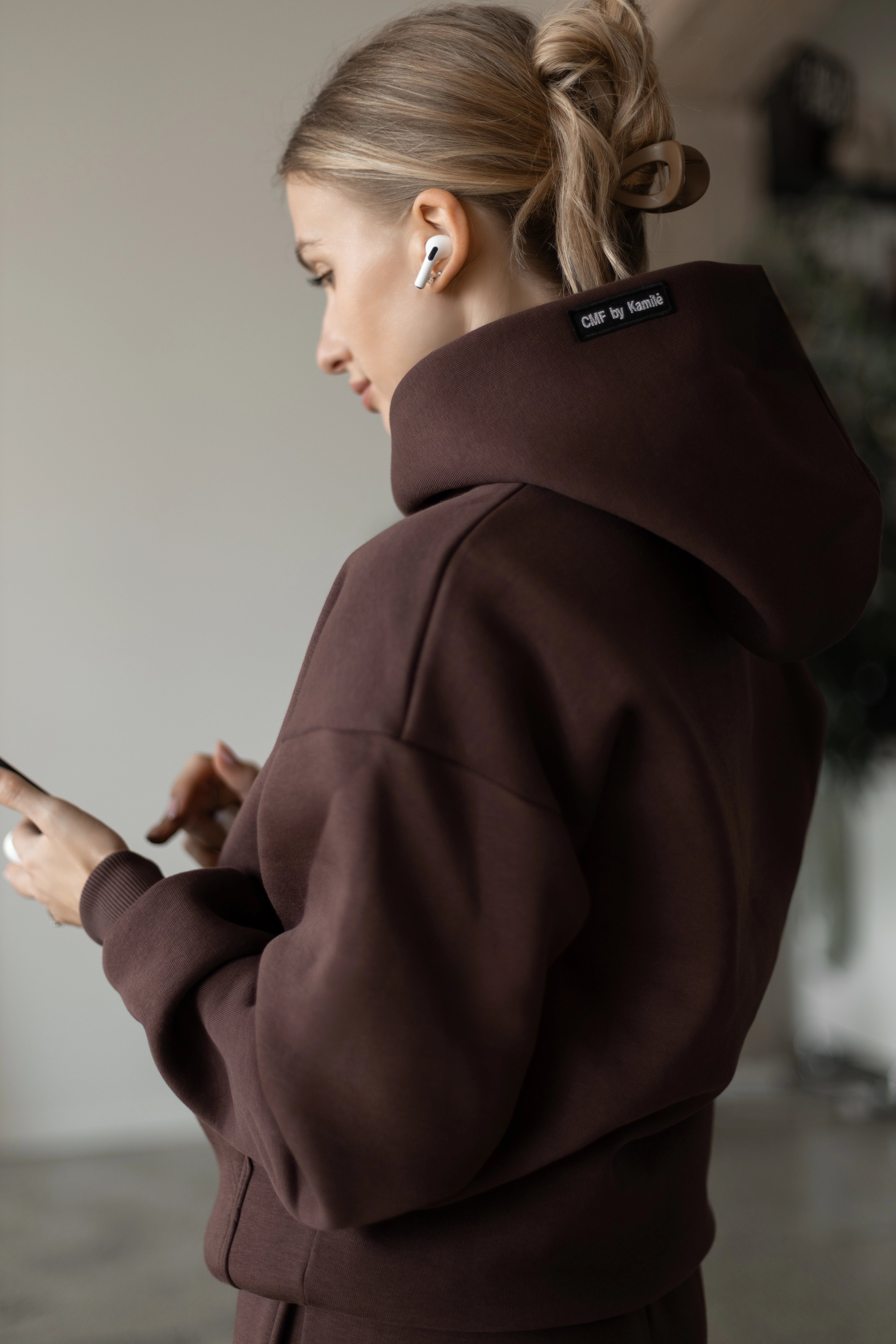 Moteris iš nugaros su tamsiai rudu treningu ir ausinėmis, rankoje laiko telefoną, matosi „CMF by Kamilė“ etiketė ant gobtuvo