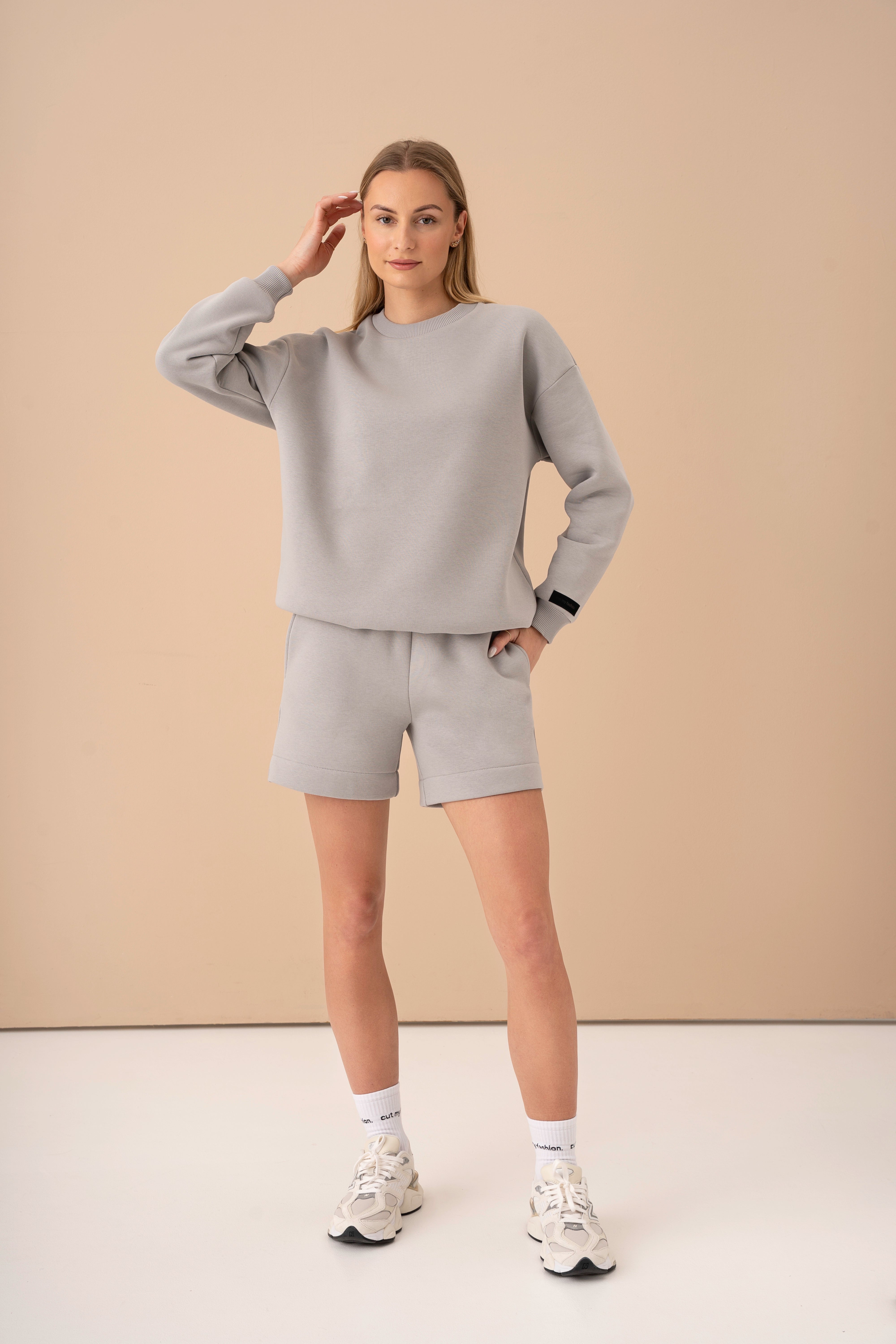 moteris su šviesiai pilku oversized cutmyfashion laisvalaikio treningu „Toma“, stovinti studijoje, ranką laiko prie veido, laisvalaikio treningas iš trisiūlio trikotažo su pūkeliu, pagamintas Lietuvoje