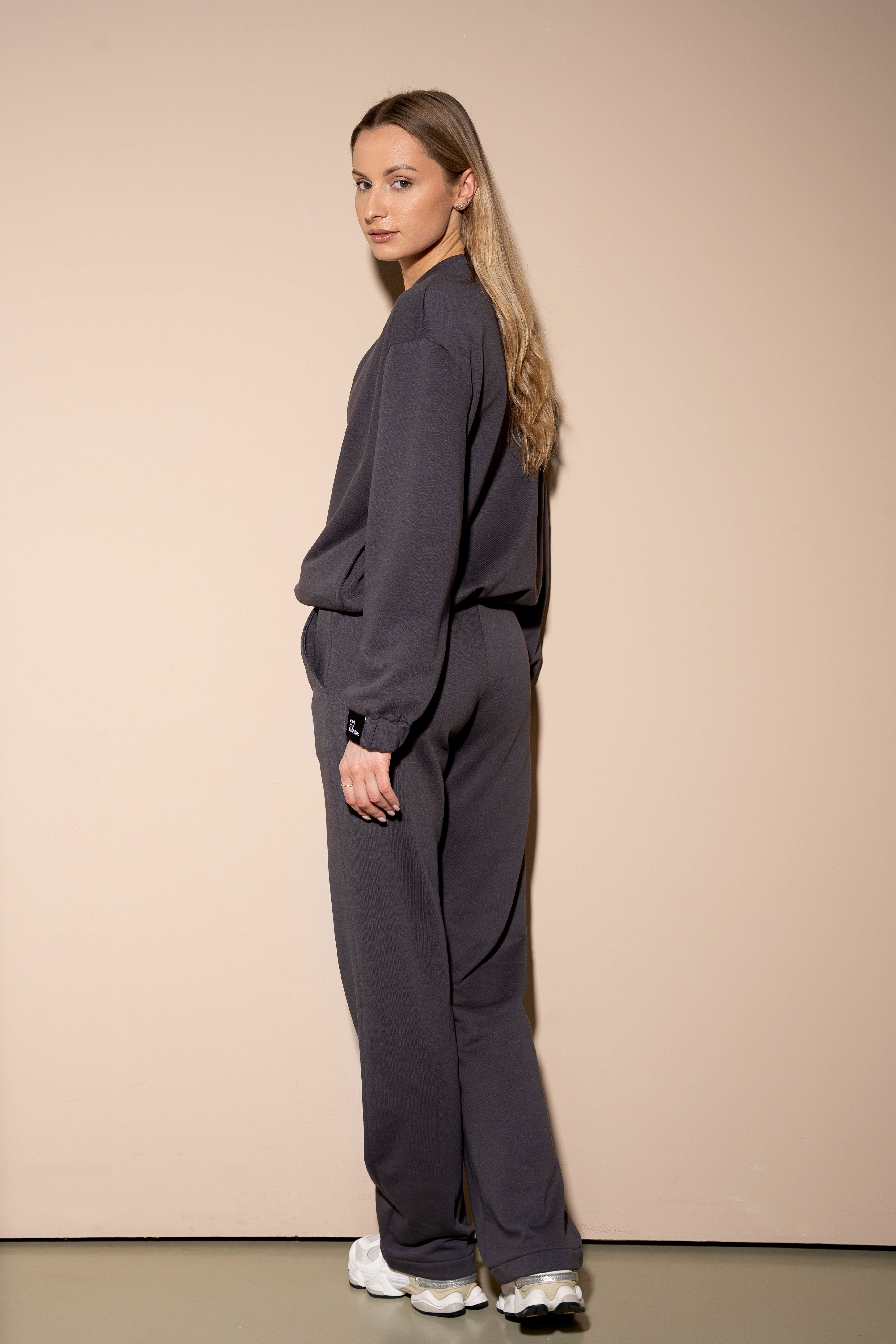 moteris dėvi tamsius oversized treningus cutmyfashion, atsisukusi šonu, žiūri atgal, studijos fone