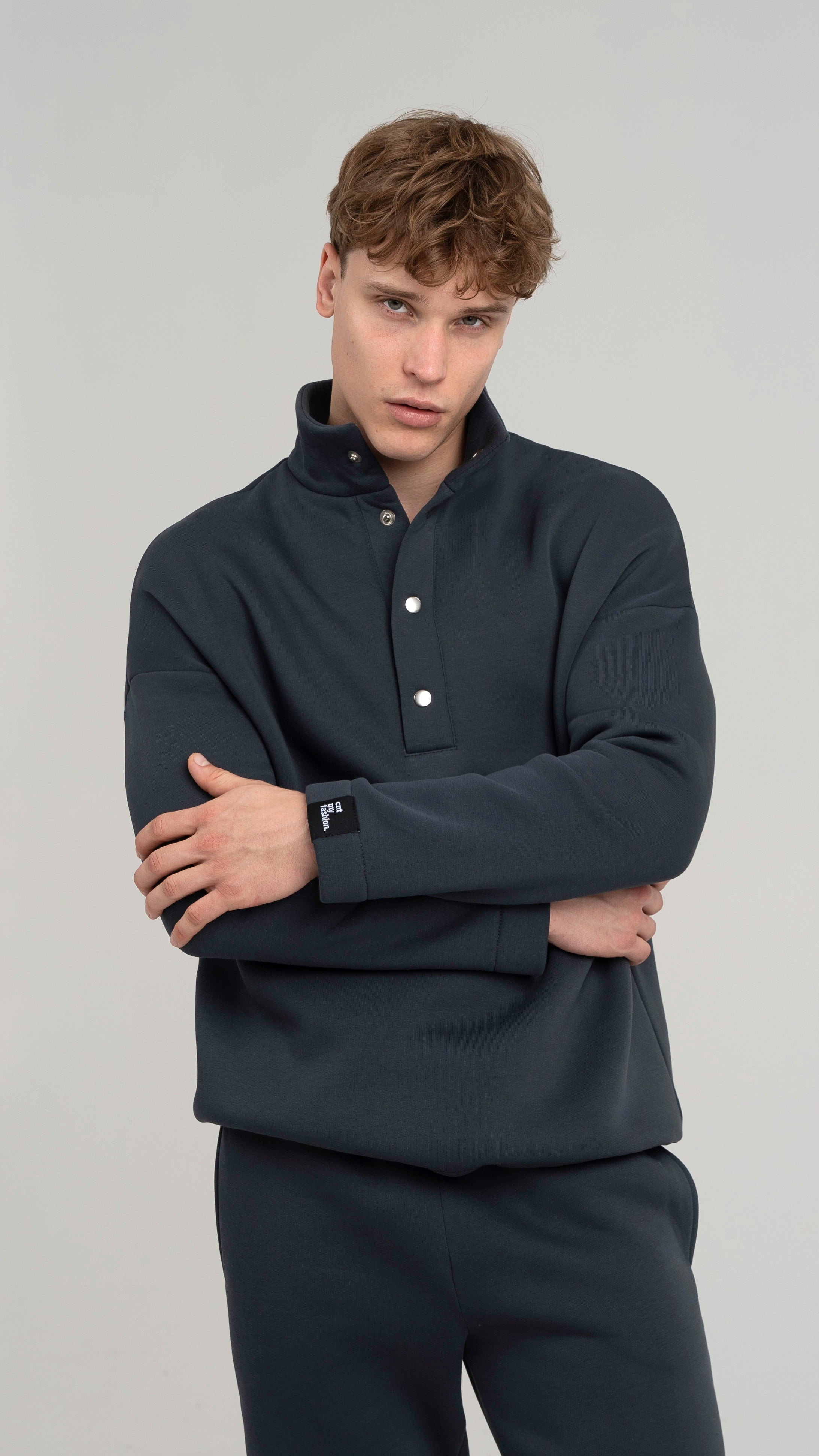 Stambus planas: vyras stovi su tamsiai pilkais treningais, su sagomis ties apykakle ir šoninėmis kišenėmis, žvelgiantis į priekį